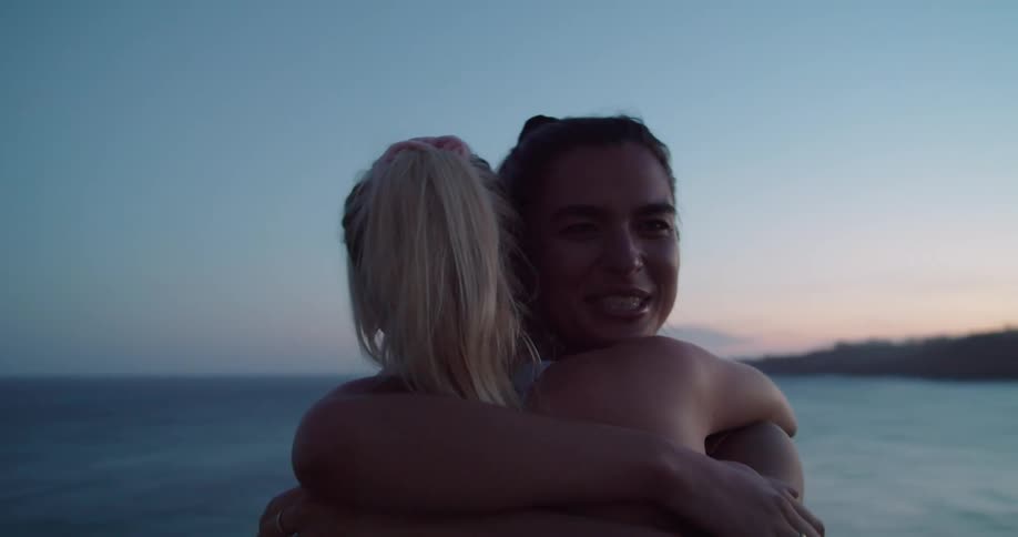 Two women hug and smile outside next to the ocean at sunset