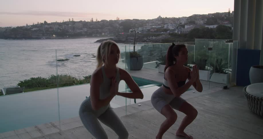 A pair of women do squats on a balcony with their backs facing the bay in the evening