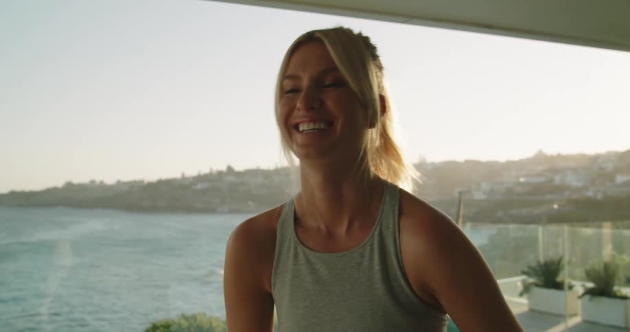 A woman tosses a workout ball up and then catches it while standing on a balcony in a beach house at sunrise