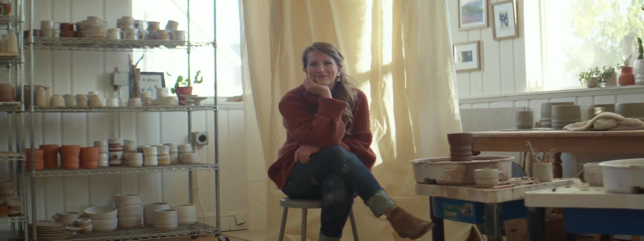 Young blonde woman wearing red sweater looks at the camera sitting in a stool in her pottery studio