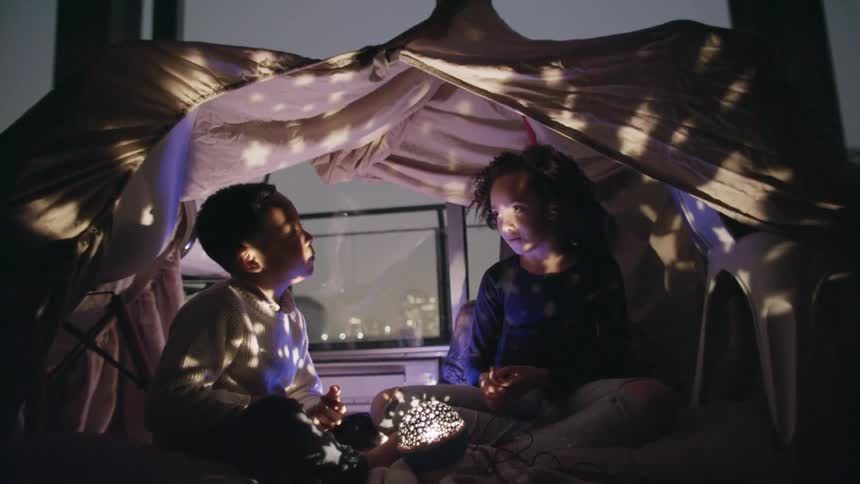 A little boy and girl sit in a tent with a light and talk in their home