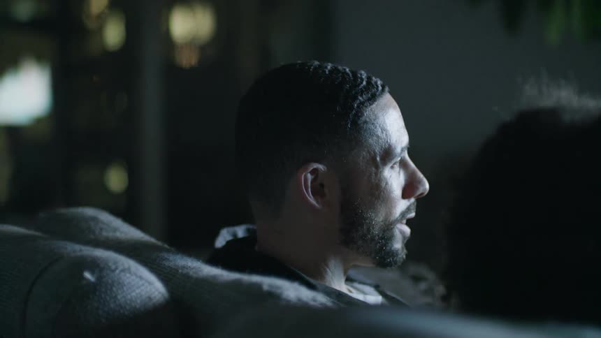 Man sitting in a couch at home talks with his partner while watching television at night