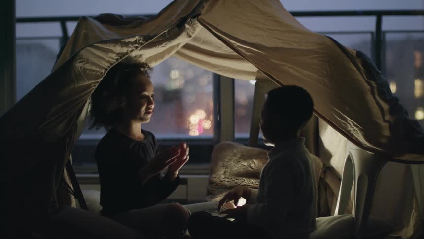 Two kids play a slap hand game sitting in a tent inside at night