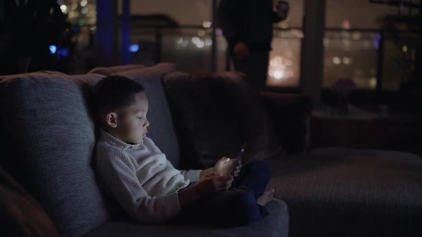 A boy sits on the couch and uses a tablet as his father stands nearby looking out a window and drinking beer