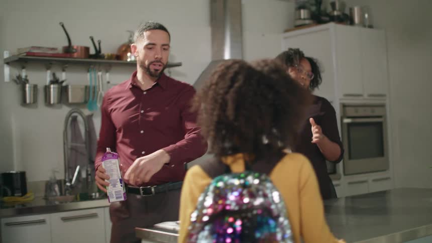 A mother and father talk to their daughter and son as they put milk and cereal away and wipe the counter