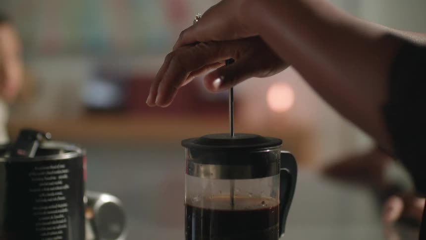 A woman makes coffee in a coffee press as someone waits by the counter