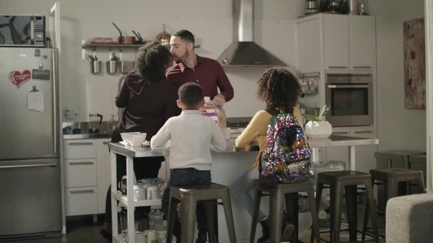 A wife reaches over to kiss her husband while they prepare breakfast for their children sitting at a kitchen island
