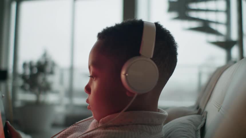 A young boy sits at home with headphones on and uses a tablet