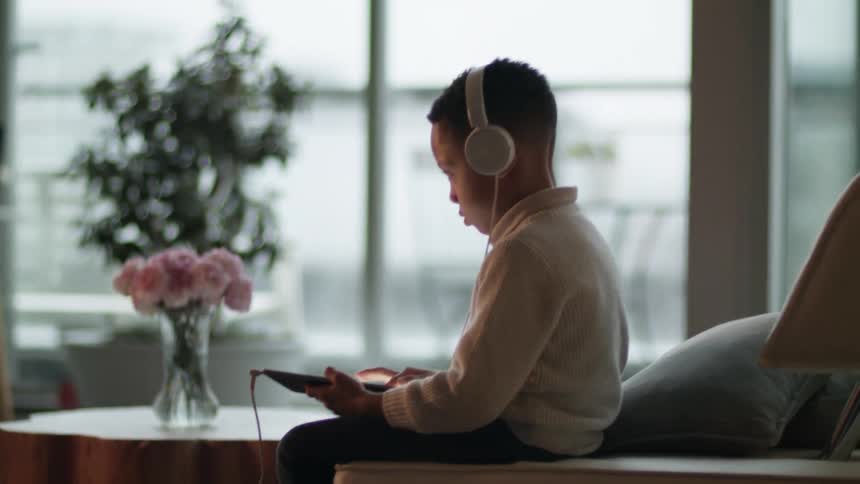 A young boy wearing a pair of headphones playing on a tablet sitting in a living room during the day