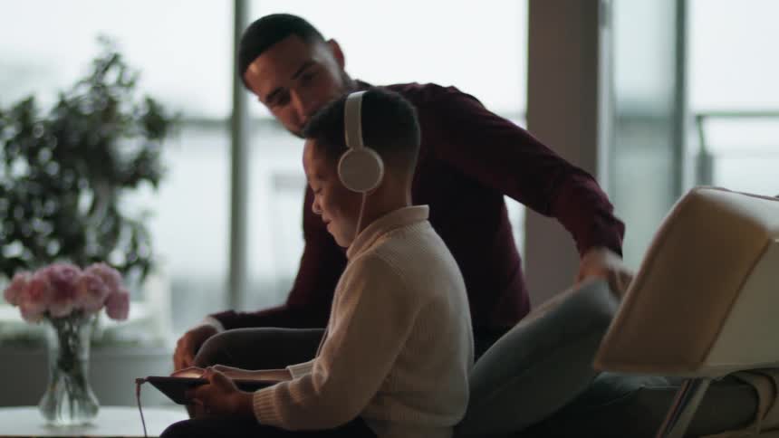 A father sitting down next to his son that is listening to music on headphones