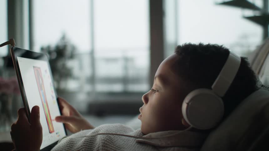 A young boy wearing headphones while using a tablet on a living room sofa