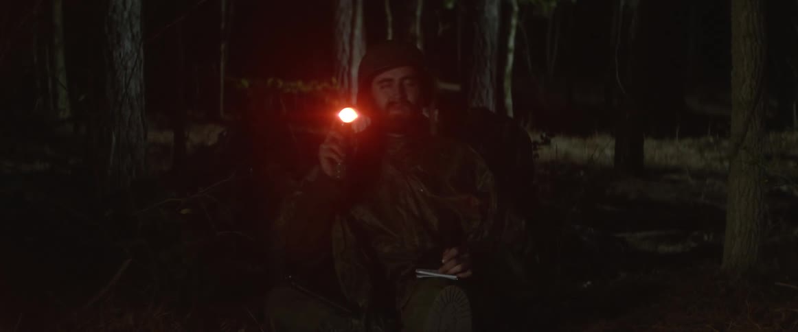 A solider sitting and writing in a forest at night with a flashlight then directed the flashlight up and talking