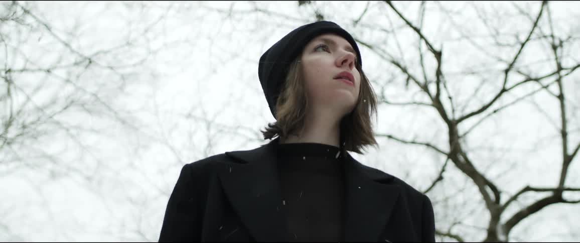 Woman wearing a black knit hat and a black coat is looking around as snow falls and trees are behind her