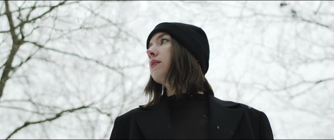 Woman wearing a black knit hat and a black coat is looking around as snow falls and trees are behind her