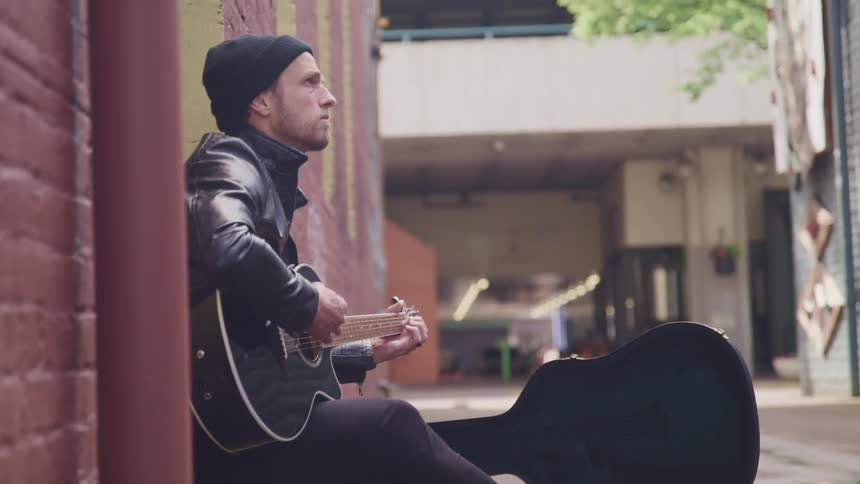 A man plays the guitar while sticking outdoors against the wall of a brick building in a city during the day