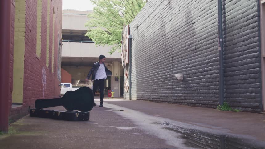 A man walking across an alley with a guitar lying in in turns around and throws a book at a brick wall