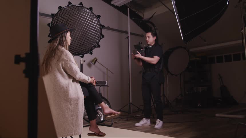 A man photographs a model as she poses on a stool in a long cardigan and hat
