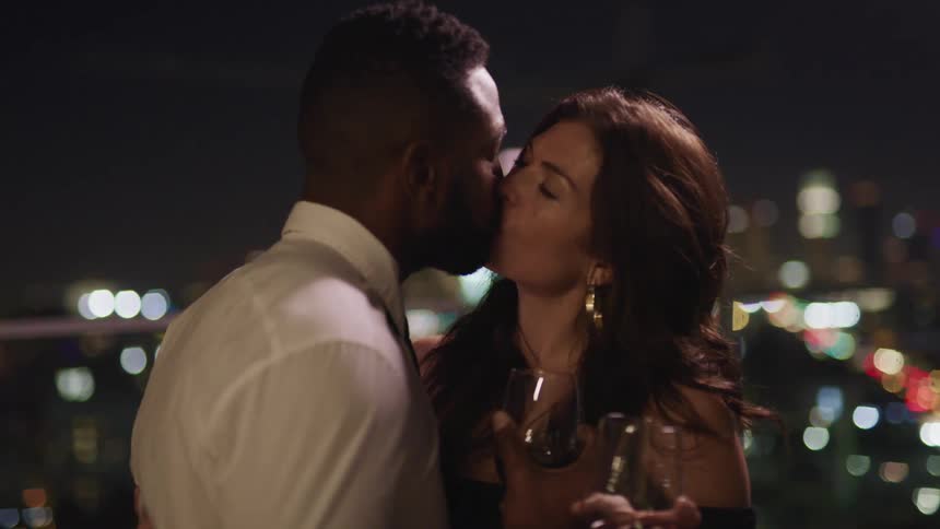 A man and woman hold wine glasses and kiss standing on top of a city building at night