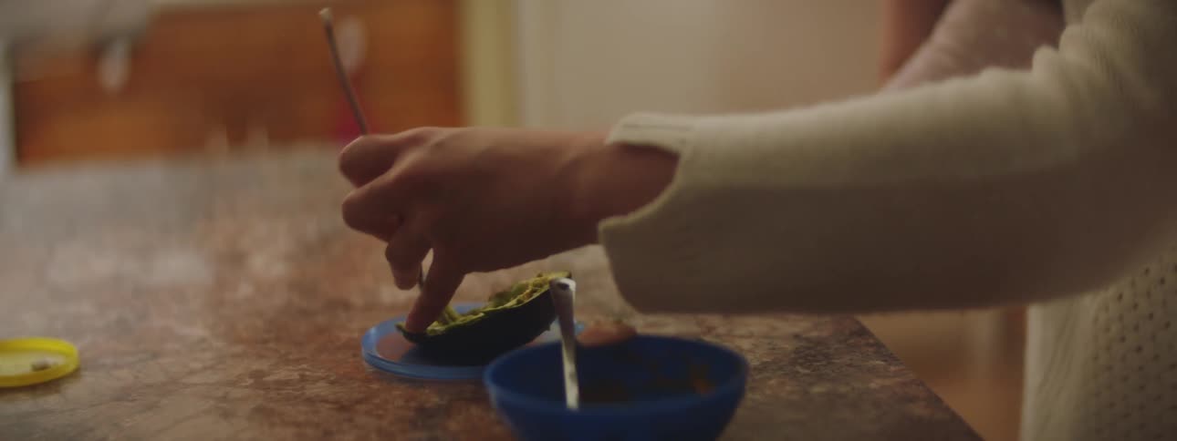 A woman at home breaks up some avocado as she holds her baby