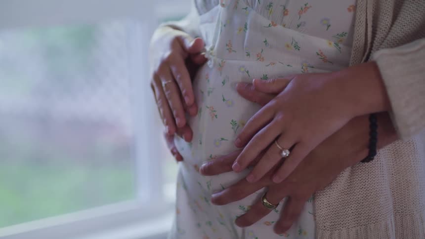 A man hugging a woman and rubbing her pregnant belly with a window in the background