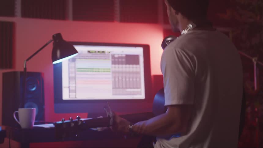 A man with a guitar and headphones in a studio sitting in front of a computer