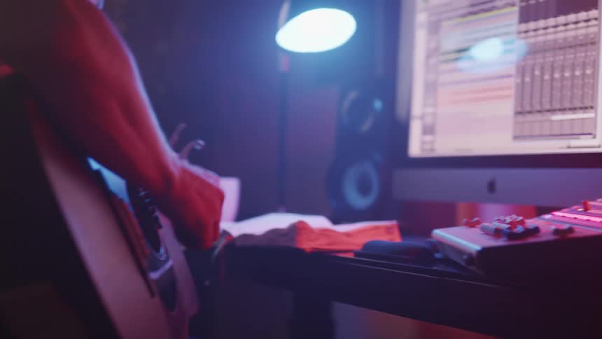A person with a guitar in a studio with a computer speakers and a lamp in the background