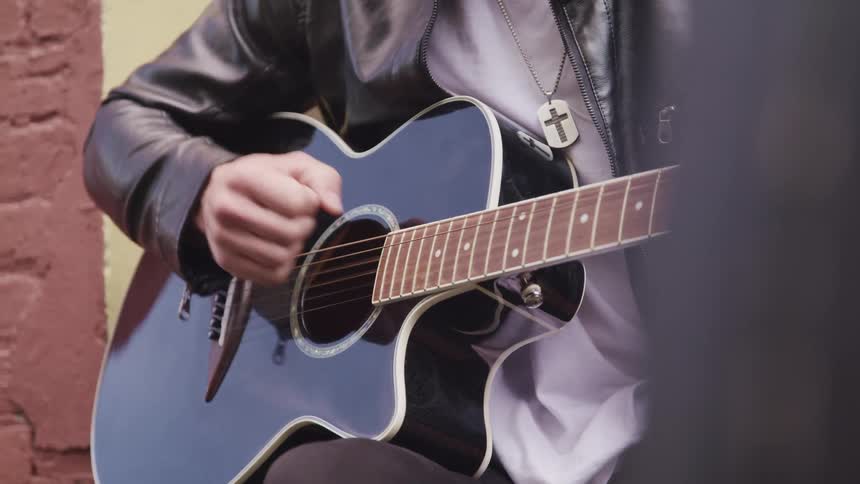 A young man taps on his guitar while sitting outside