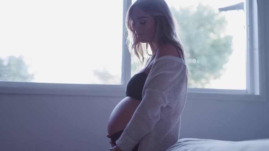 Young pregnant woman in black underwear caressing her belly over a bed at home