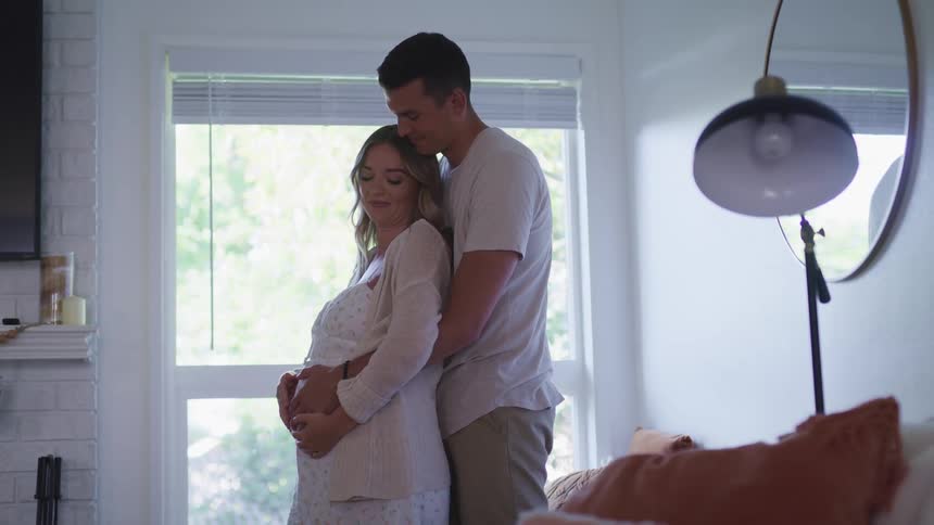 A man stands behind his wife with his arms wrapped around her and they both rub her pregnant belly