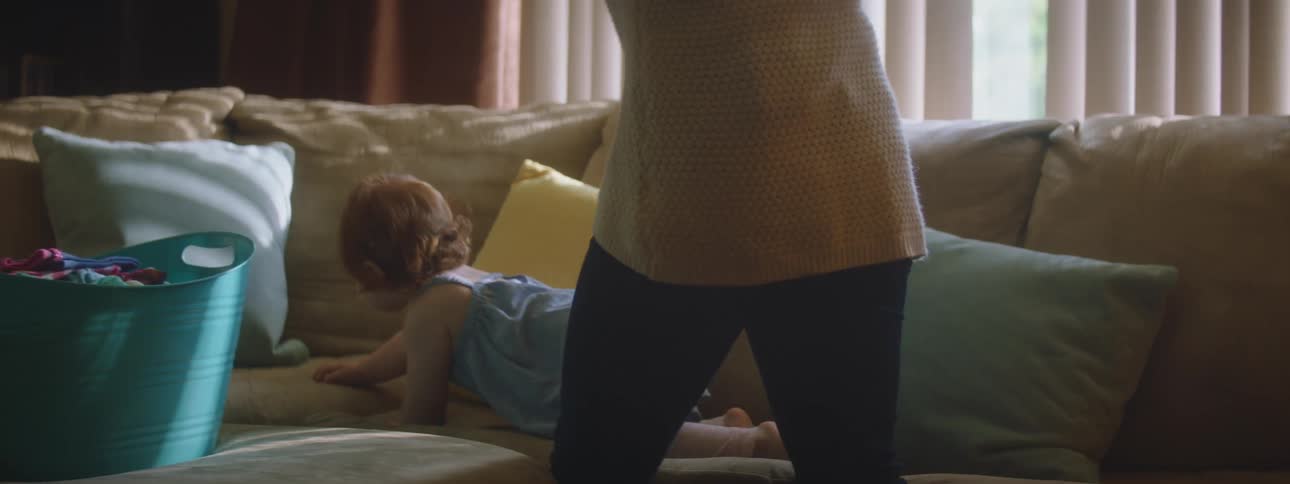 A woman in a room standing by a couch touching a baby crawling on the couch with windows and blinds in the background