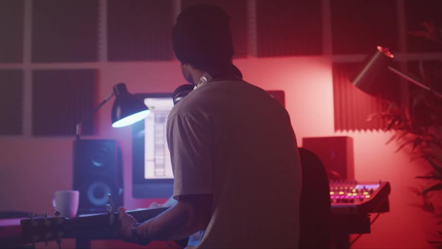 A man at home sits down in front of his computer with his guitar and starts playing