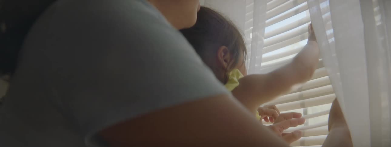 A woman is holding a little girl near a window while they play with the blinds