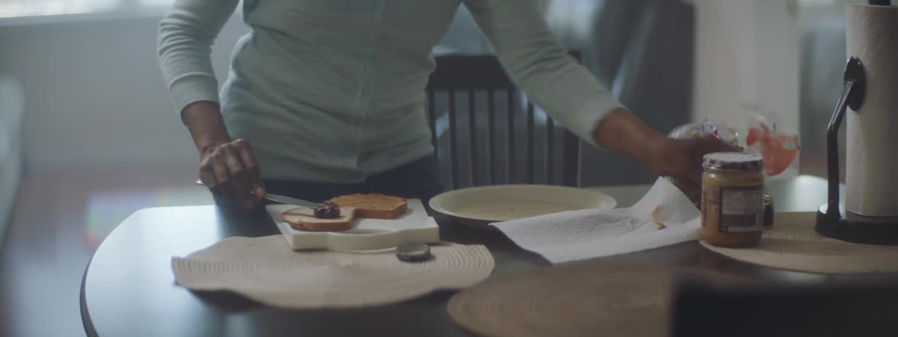 A woman is making a peanut butter and jelly sandwich on a cutting board on a kitchen table
