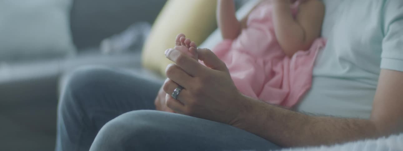 A father rubbing an infant daughters bare foot while sitting on a sofa