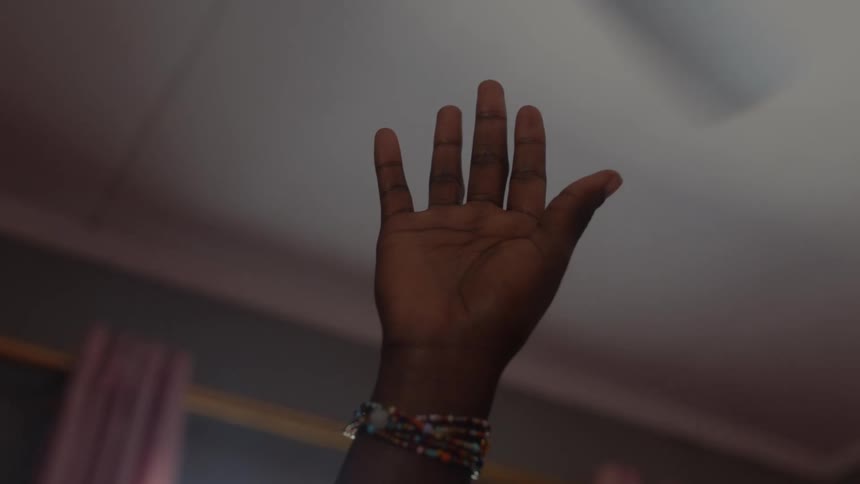 A person in a room with their hand up with a ceiling fan above and a window and curtains in the background
