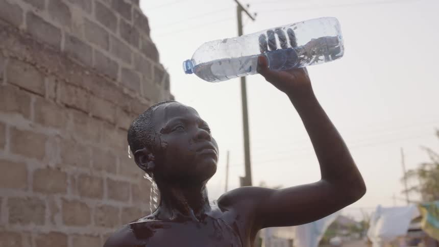 A young boy stands outdoors and pours a bottle of water over his head to cool off