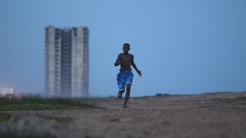 A shirtless boy sprinting across dirt with a tall building in the distance