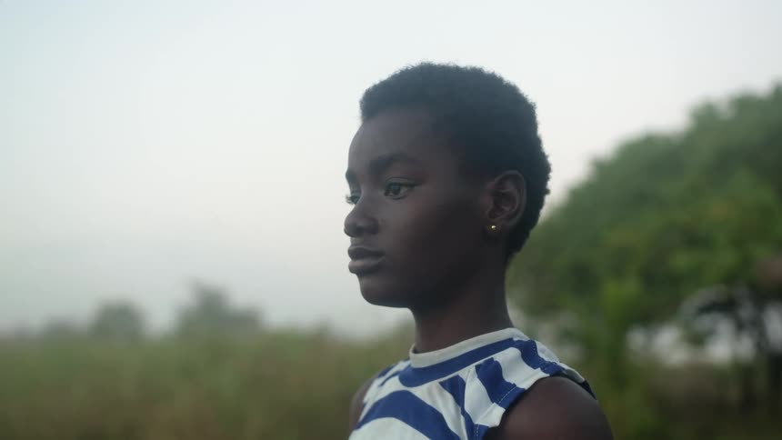 A young girl standing on the shore rubs her shoulder and looks out at the water