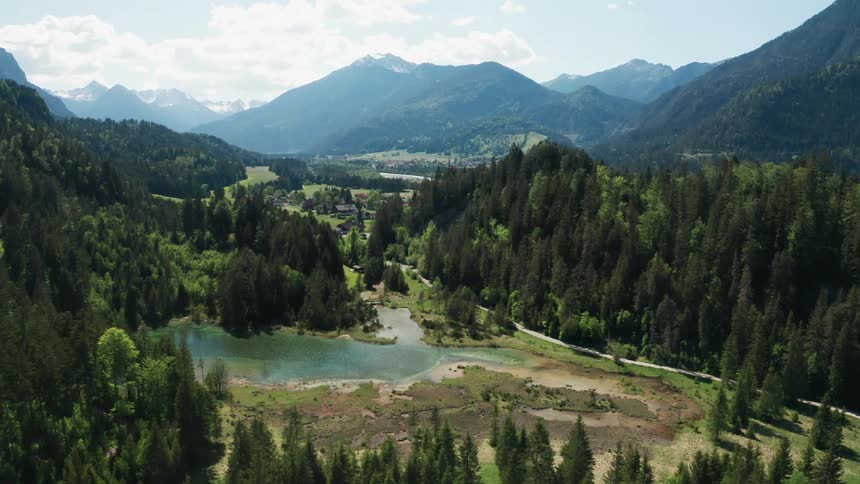 A pond surrounded by trees next to a road with a mountain range the sky and clouds in the background