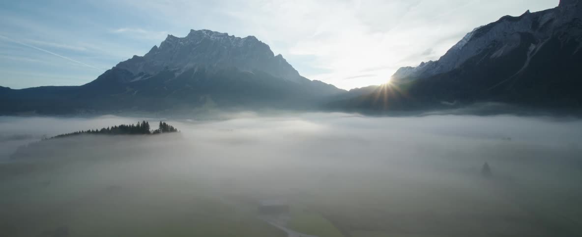A thick layer of fog covers the valley below a tall mountain range as the sun peeks over the mountains