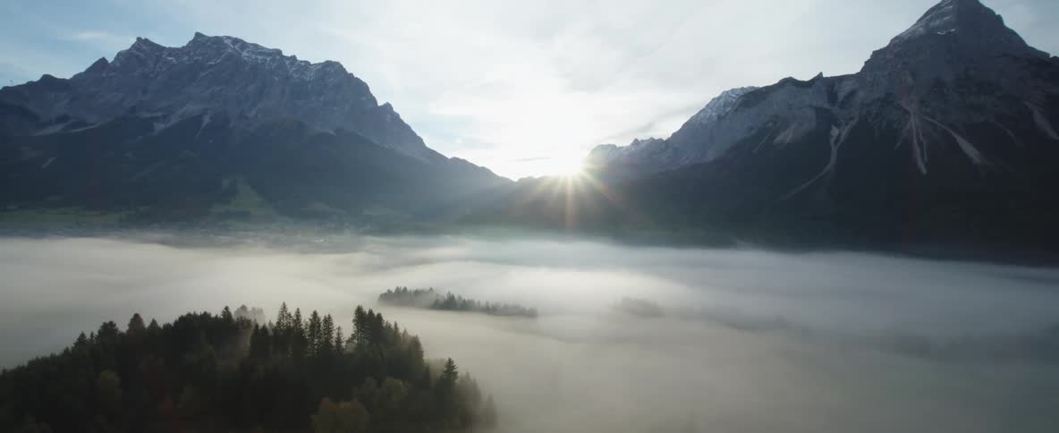 Aerial view of white fog covering the pine tree forests at the foot of the snow covered peaks of a mountain range at sunset