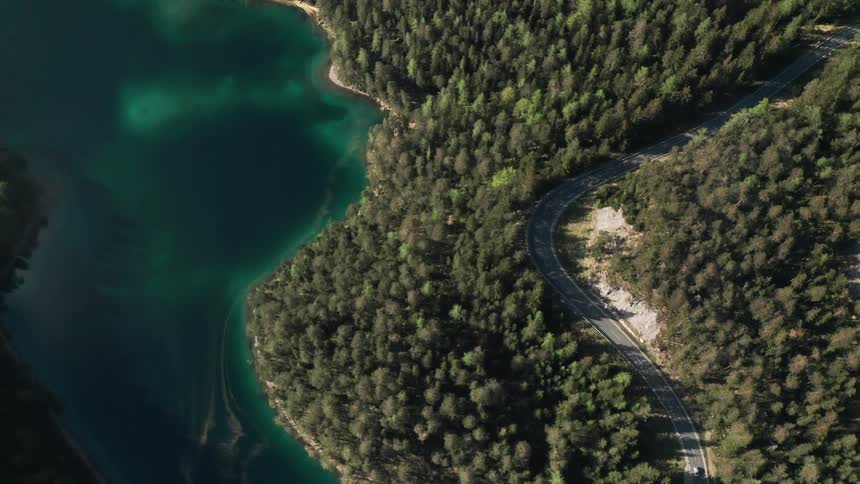 Vehicles travel along a winding road through a forest bordering a river on a sunny day