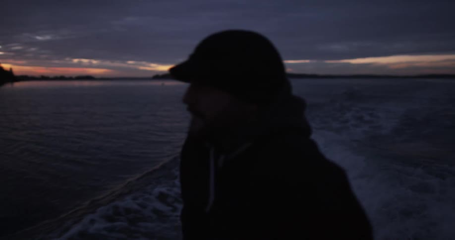 A man looks off to the side of a boat moving across water after sunset