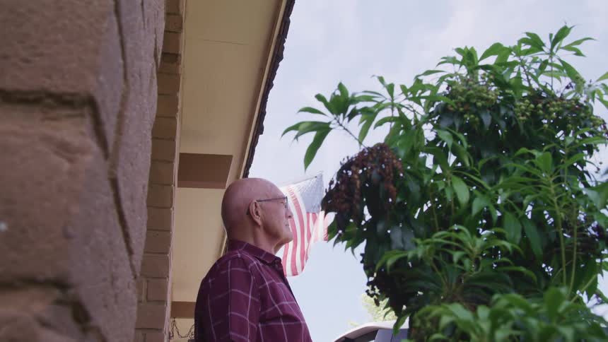 A man standing in a front porch with an american flag the sky and clouds in the background