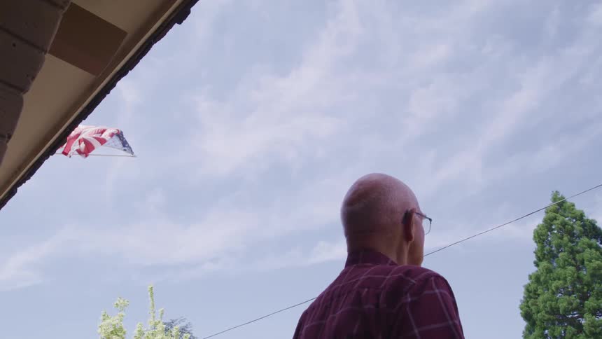 A man stands outside his home as a flag above him blows in the wind