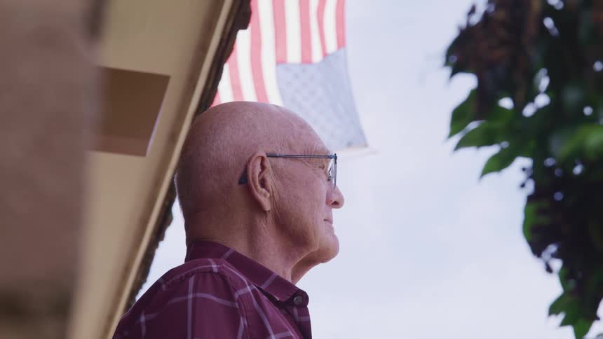 A man standing in front of a house with the american flag the sky and clouds in the background