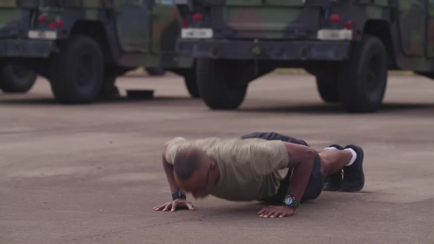 A man with glasses doing push ups in a military base with vehicles in the background