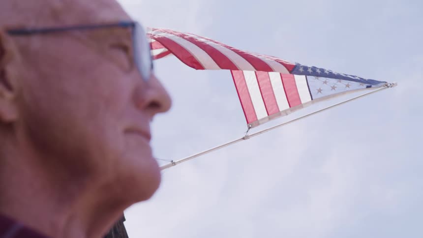 A man with glasses standing with the american flag the sky and clouds in the background