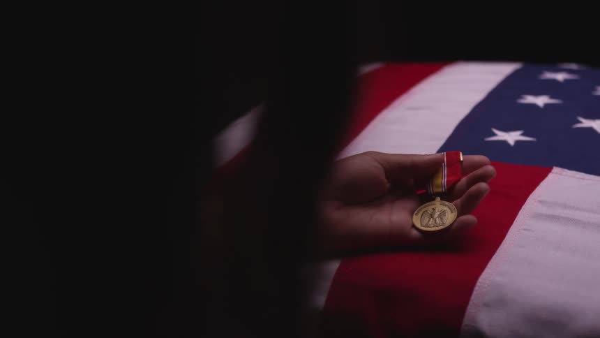 A persons hand resting on a casket with an american flag holding a soldiers medal