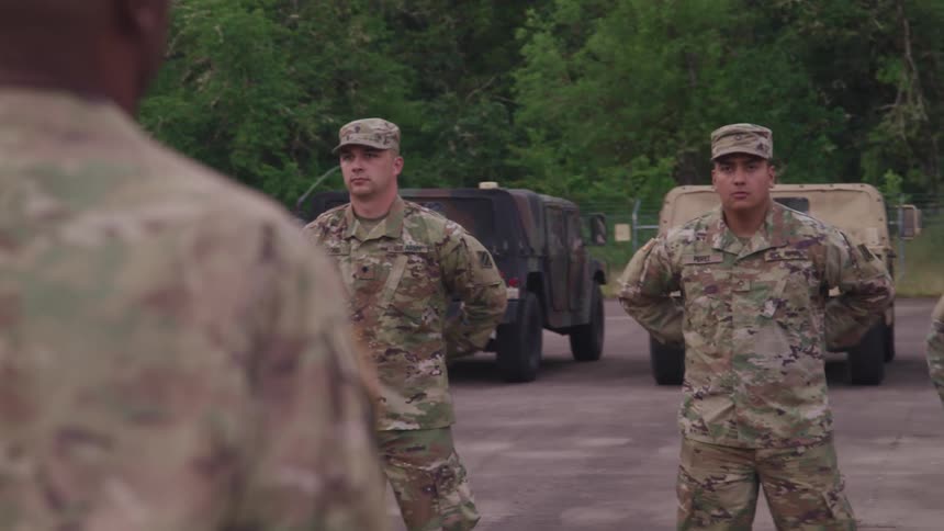 Drill instructor ordering a troop of american soldiers to stand at ease on a yard near vehicles